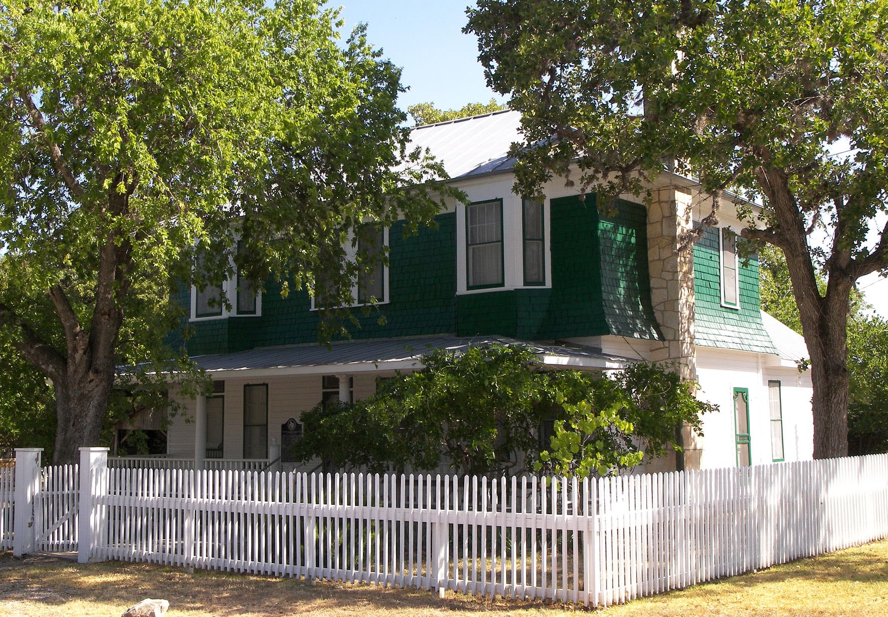 A white picket fence and green house in the middle of a yard.